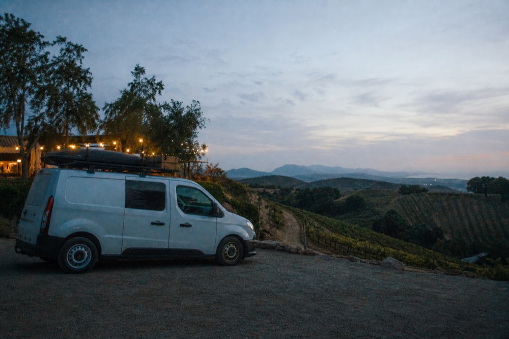A small touring van parked at a hilltop vineyard pullout with vine rows falling away toward distant hills, no text, no logos, no people