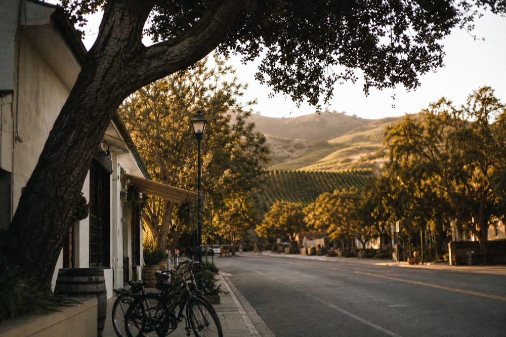 A quiet small-town main street with a few storefronts and parked bicycles under oak trees in golden afternoon light, no text, no logos, no people