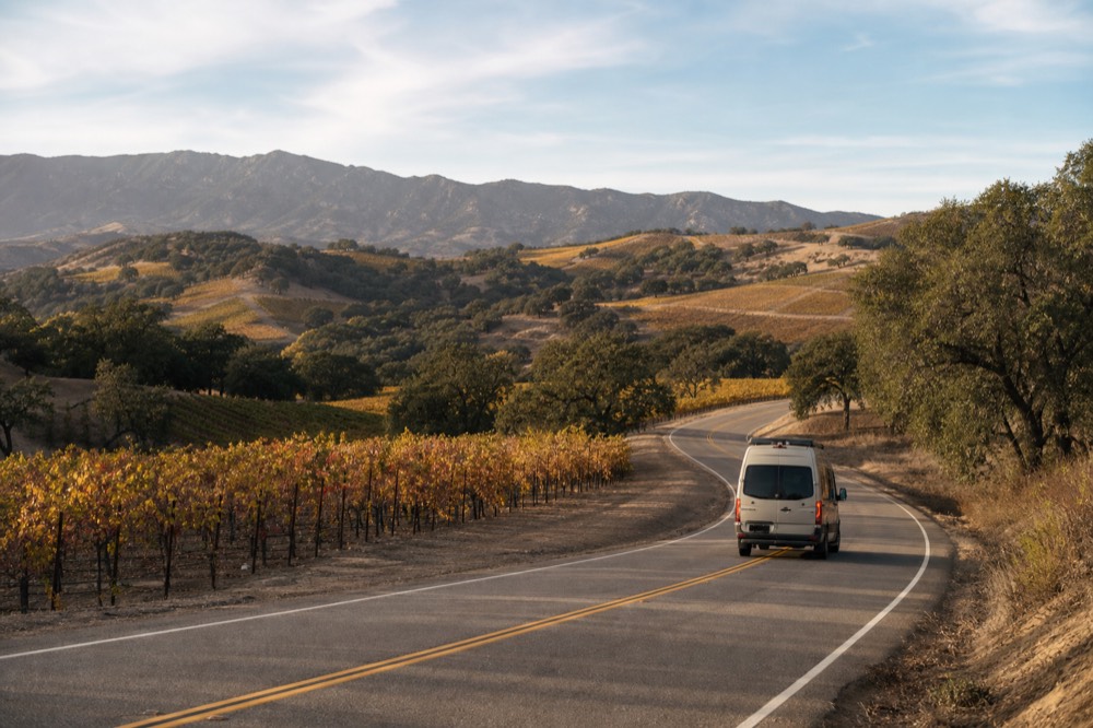 A small touring van on a two-lane valley road winding between vineyard blocks and oak trees under a wide afternoon sky, no text, no logos, no people