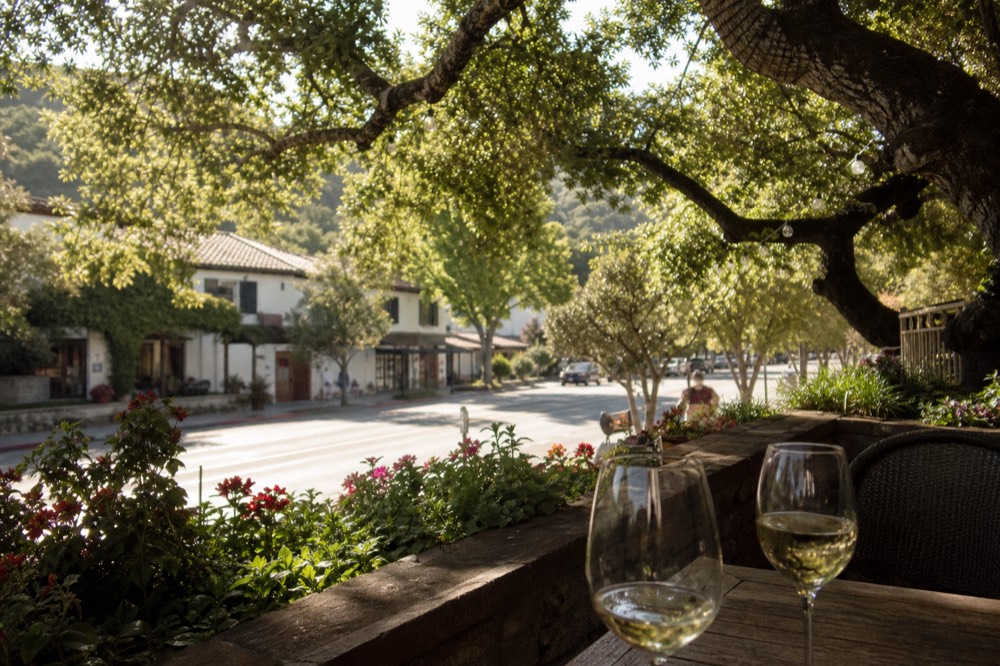 Two wine glasses on an outdoor table on a flower-lined village patio with a quiet main street beyond in warm afternoon light, no text, no logos, no people