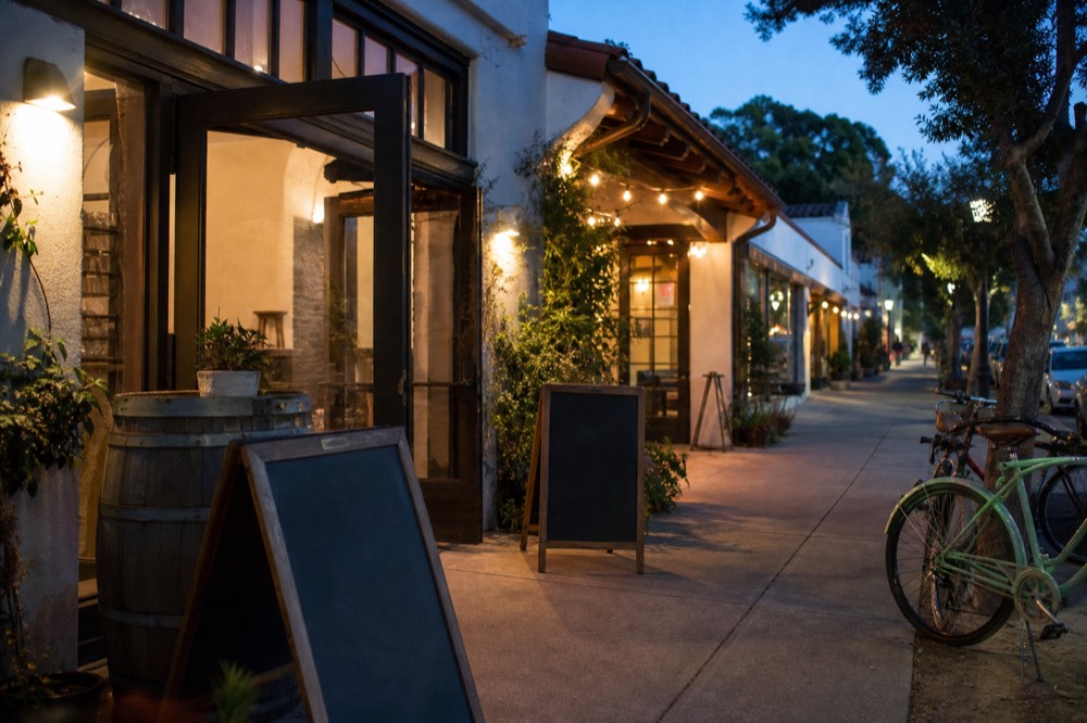 A relaxed walkable small-town street of tasting-room storefronts with open doors, sidewalk chalkboards blank and a couple of parked bicycles, warm light, no text, no logos, no people