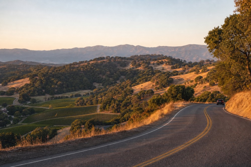 A two-lane road climbing over a wooded mountain pass with oak-studded golden hills and a glimpse of vineyards far below, no text, no logos, no people