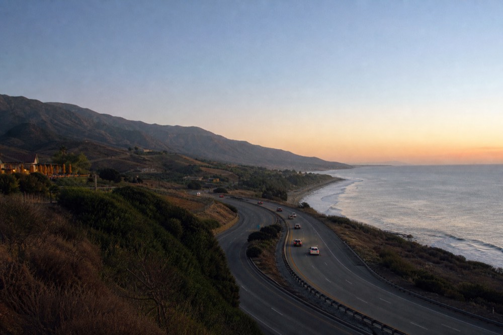 A coastal freeway curving along the shoreline with the Pacific on one side and golden hills on the other in bright daylight, no text, no logos, no people