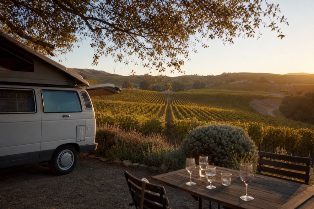 A small touring van parked at a vineyard estate with guests' glasses on an outdoor table and rows of vines stretching behind under golden light, no text, no logos, no people
