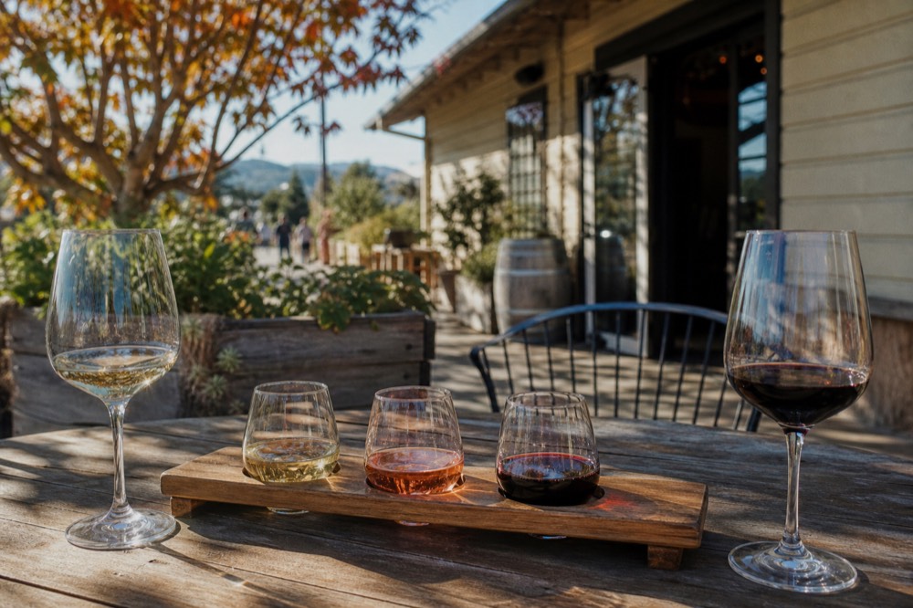 Two people's wine glasses beside a single shared tasting flight on a sunny sidewalk table outside a small-town tasting room, no text, no logos, no people