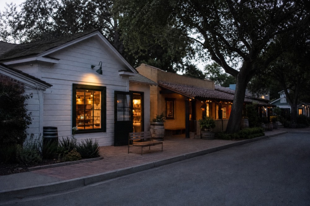 A cluster of small clapboard and adobe tasting-room storefronts along a quiet village street shaded by oak trees, no text, no logos, no people