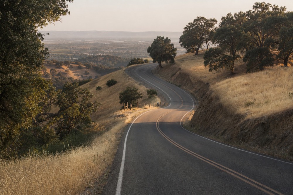 A two-lane road climbing over the oak-studded San Marcos Pass with golden hills and a distant view of the inland valley, no text, no logos, no people