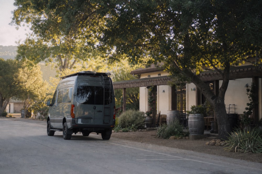 A small touring van parked on a quiet village street beside tasting-room storefronts under oak trees in late afternoon light, no text, no logos, no people