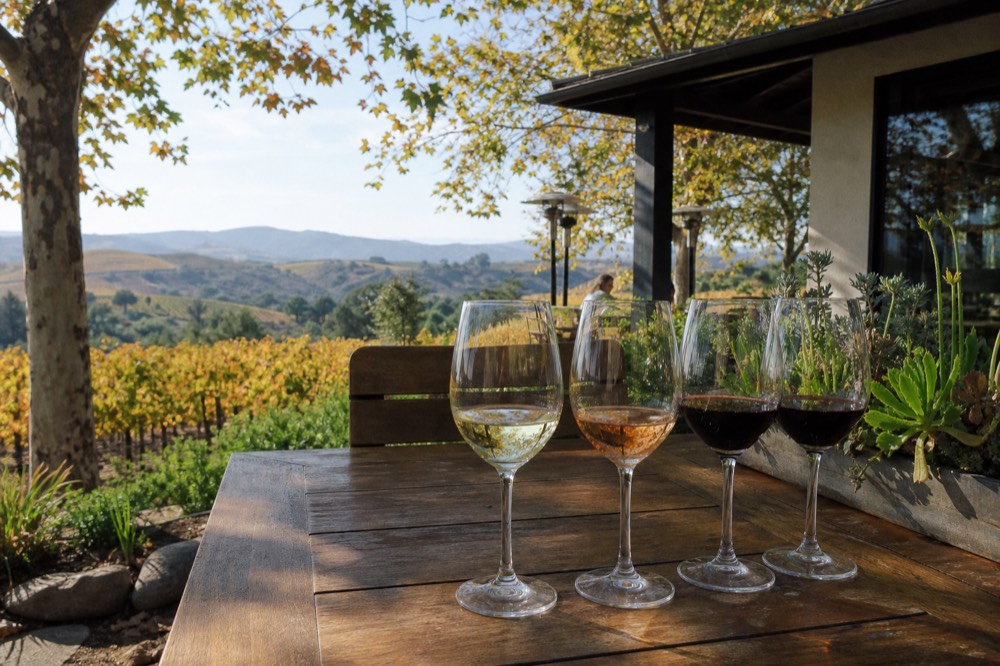 Wine glasses catching afternoon light on a wooden table at a Solvang tasting patio, vineyard hills soft in the distance, no text, no logos, no people