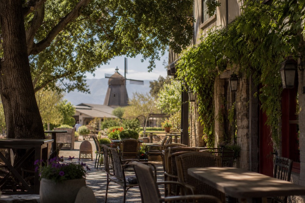 A Solvang streetscape with outdoor cafe seating, potted flowers and a wooden windmill soft in the background, warm midday light, no text, no logos, no people