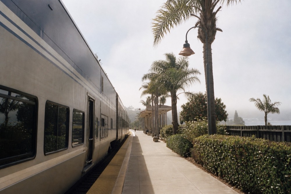 A modern coastal passenger train stopped at a small palm-lined station platform near a downtown waterfront, no text, no logos, no people