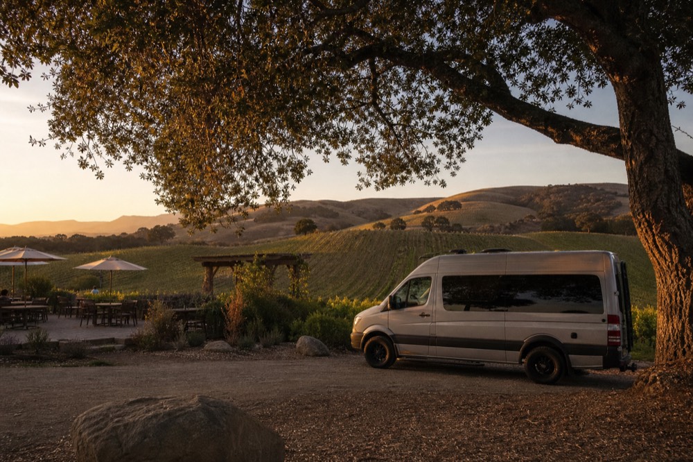 A small touring van parked beside a vineyard tasting patio with oak trees and rolling vine rows behind, no text, no logos, no people