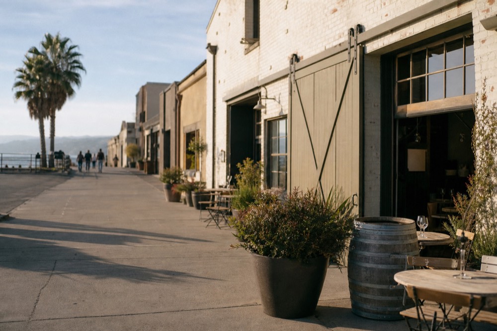 A sunlit coastal warehouse-district tasting street with roll-up doors, potted plants and bistro tables near the waterfront, no text, no logos, no people