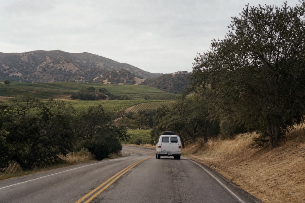 A small touring van on a two-lane valley road winding between oak trees and vineyard blocks under a wide afternoon sky, no text, no logos, no people