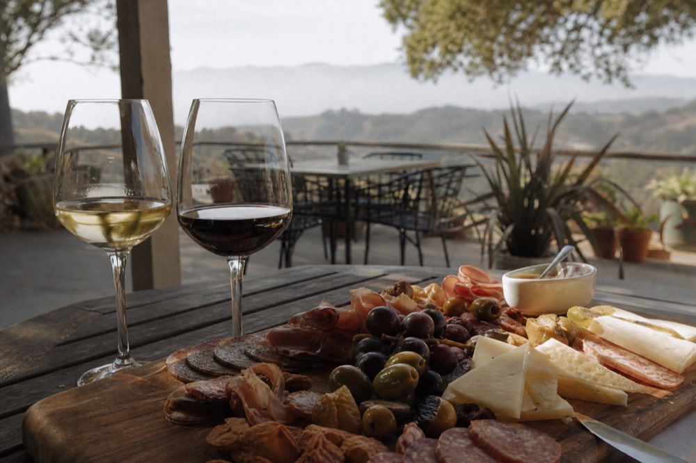 A close view of a cheese and charcuterie board with olives and grapes beside two glasses of red and white wine on a rustic table, no text, no logos, no people