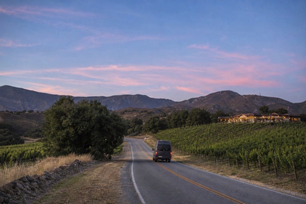 A small touring van on a two-lane valley road winding between vineyard blocks and oak trees under a wide afternoon sky, no text, no logos, no people