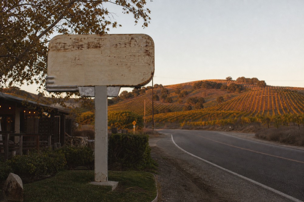 A weathered roadside restaurant sign shape at dusk beside a two-lane valley highway with vineyard hills behind, no text, no logos, no people