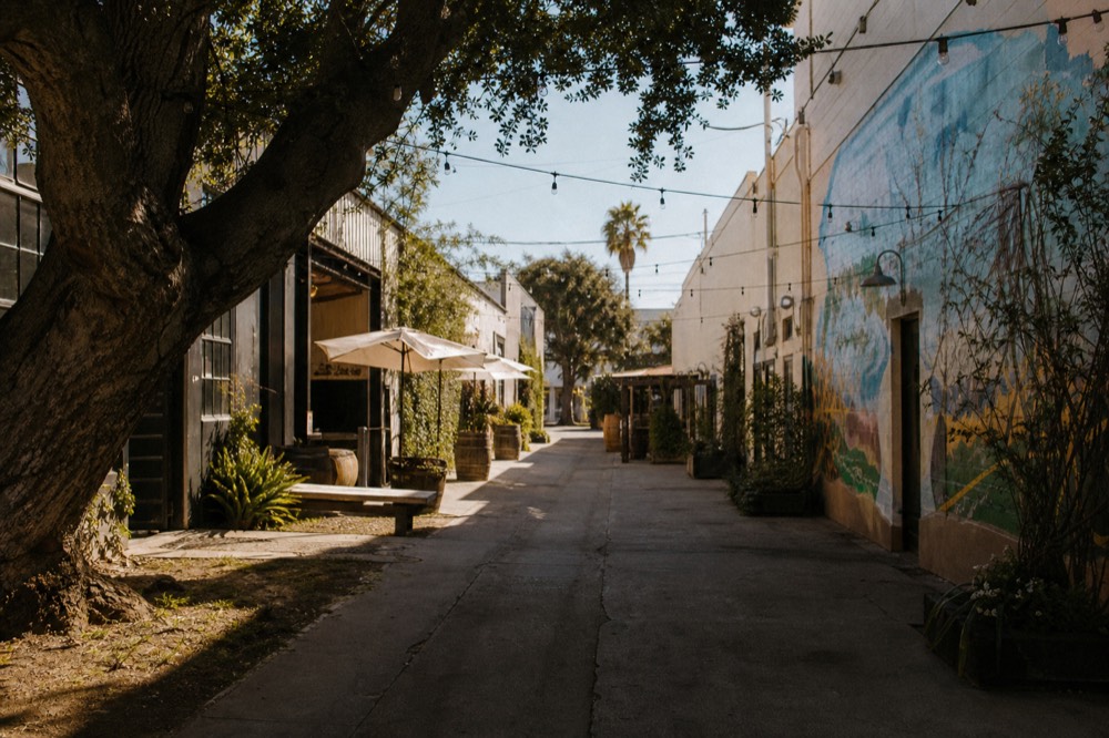 A narrow Funk Zone lane lined with converted warehouse tasting rooms and a bright wall mural, string lights overhead in late afternoon sun, no text, no logos, no people