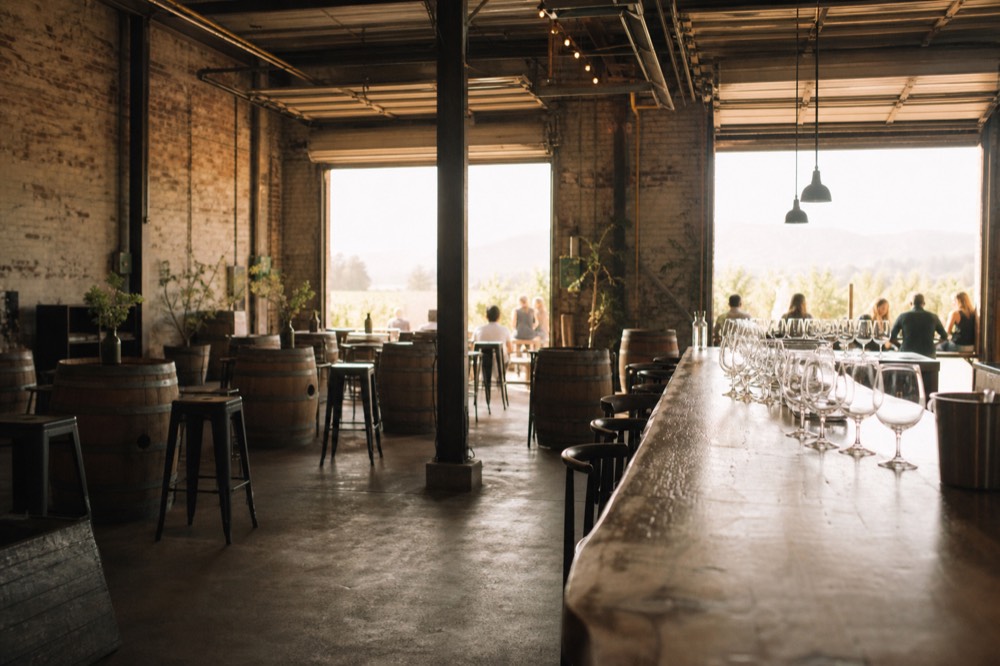 Inside a converted warehouse tasting room with exposed brick, a concrete floor and a long wooden bar lined with wine glasses, warm light from roll-up doors, no text, no logos, no people
