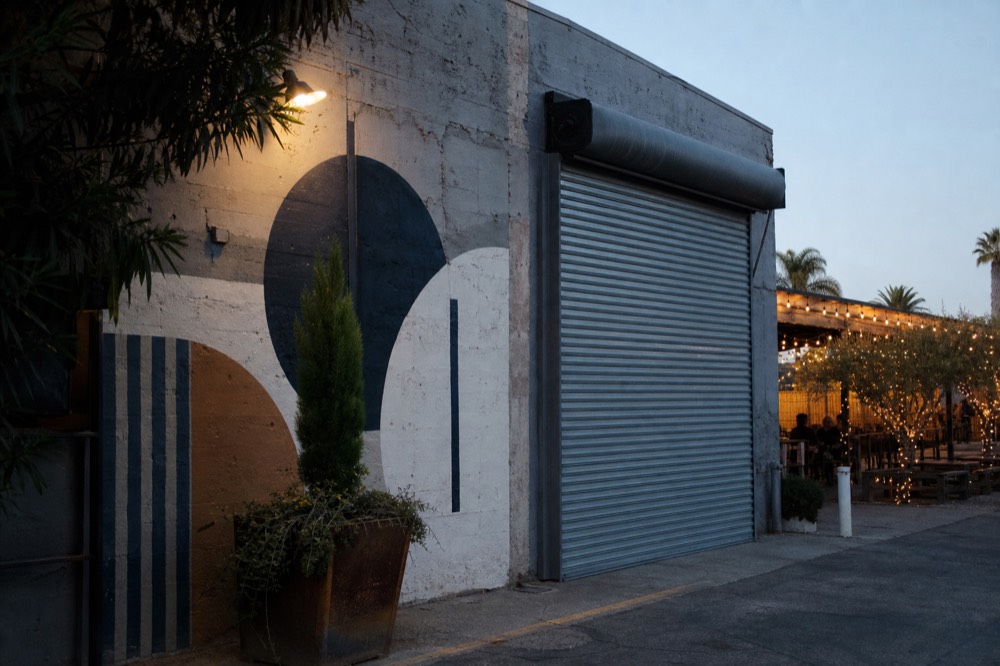 A weathered warehouse wall with a roll-up metal door and an abstract painted mural in the Funk Zone under bright afternoon light, no text, no logos, no people
