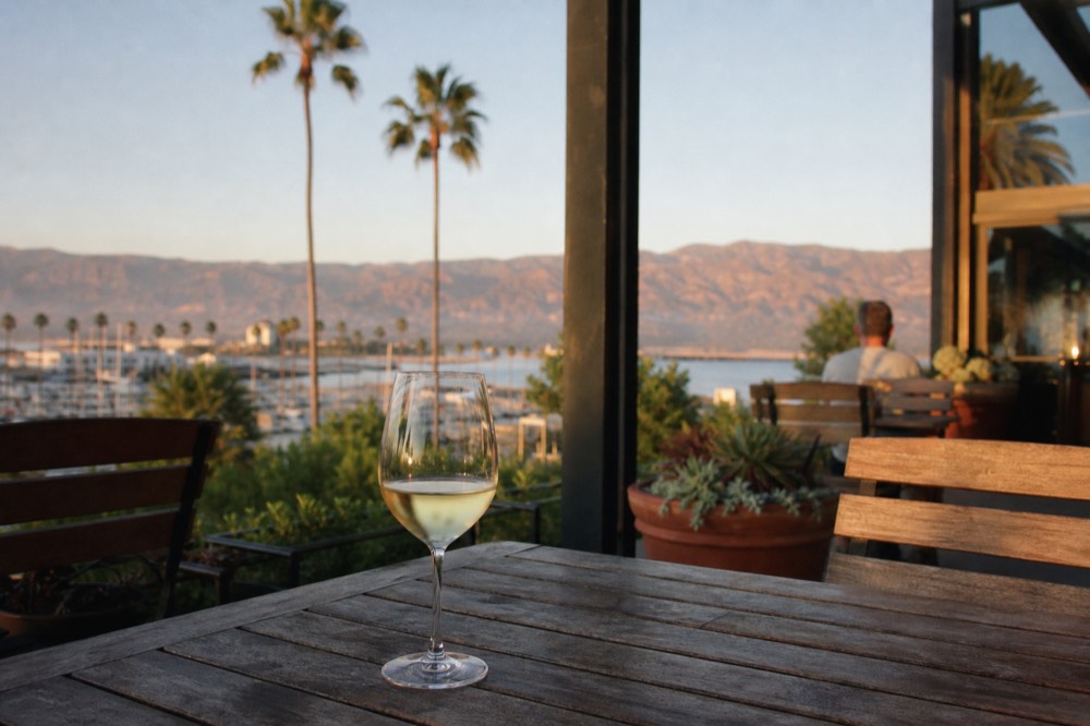 A glass of white wine on an outdoor Funk Zone patio table with the harbor and palm trees soft behind in golden-hour light, no text, no logos, no people