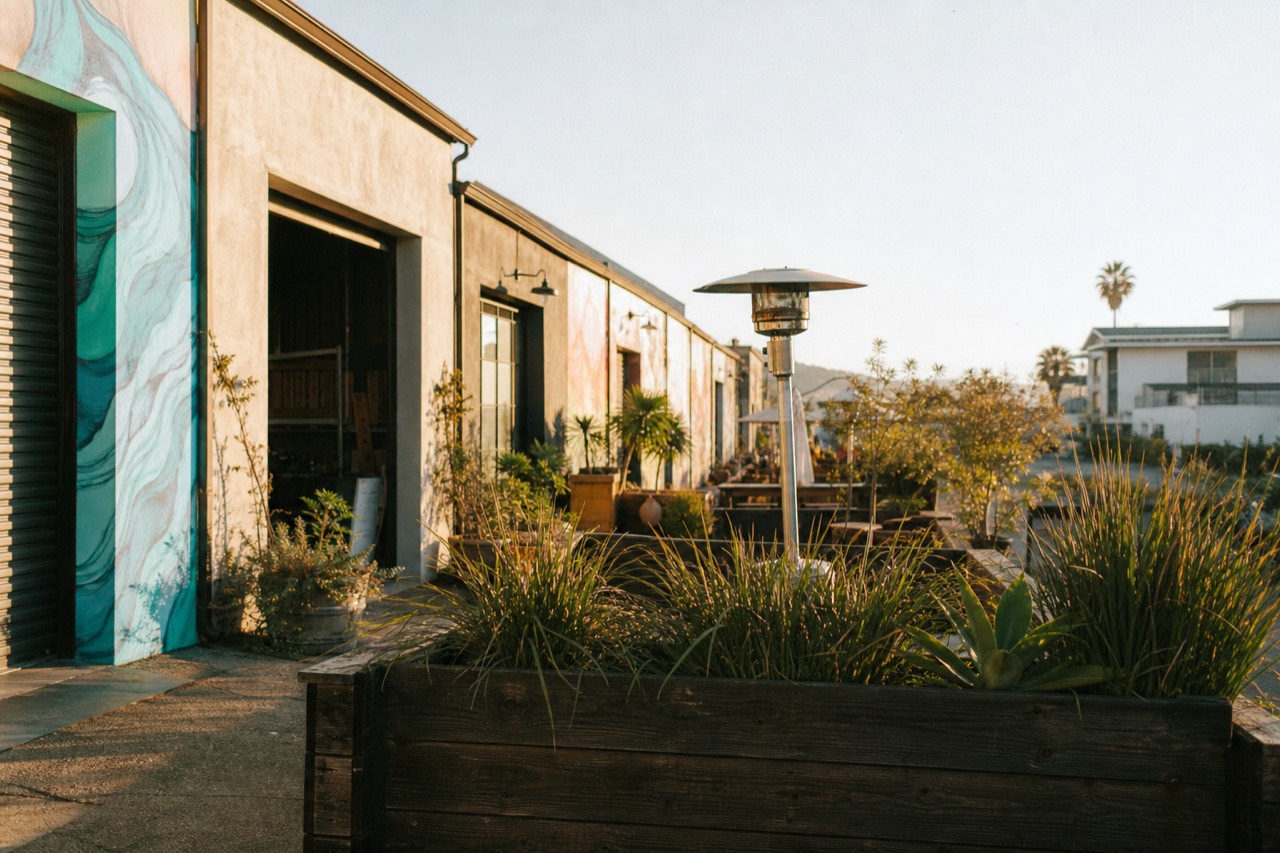 A sunny Funk Zone streetscape of low warehouse buildings converted to tasting rooms with murals and roll-up doors near the Santa Barbara waterfront, no text, no logos, no people