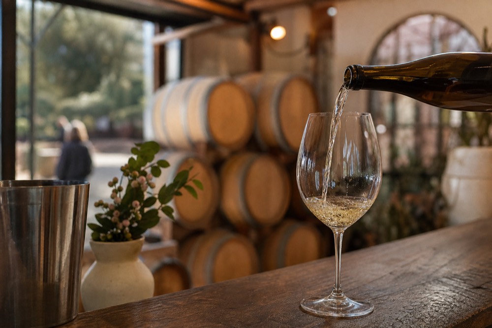A wine pour caught mid-stream into a glass at a rustic estate cellar-door counter with barrels blurred behind, no text, no logos, no people