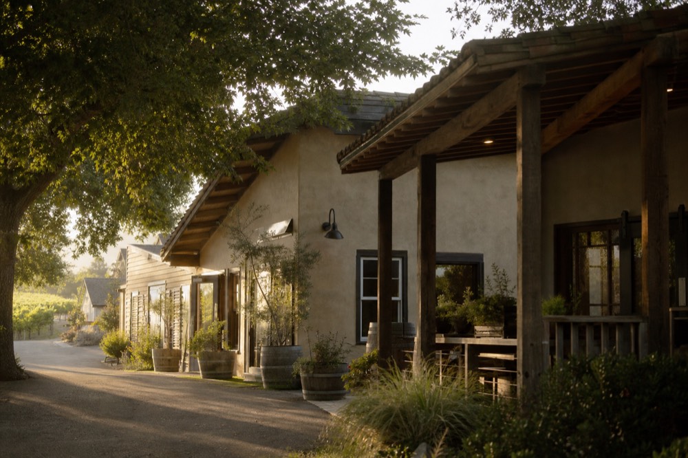 A cluster of low adobe and clapboard tasting-room storefronts along a walkable valley village street shaded by trees, no text, no logos, no people