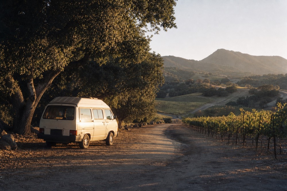 A small touring van parked beside oak-shaded vineyard rows on a rural ranch road in warm afternoon light, no text, no logos, no people