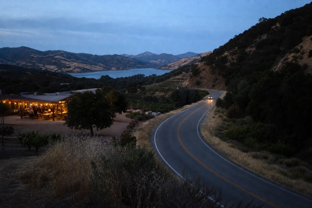 A winding mountain highway curving through chaparral and oak on the San Marcos Pass with a distant blue reservoir and golden ridgelines, no text, no logos, no people