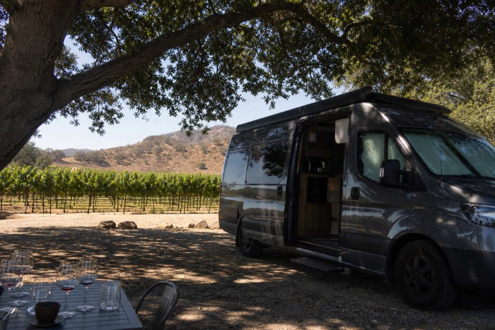 A small touring van parked beside oak-shaded vineyard rows on a valley ranch road with tasting glasses on a nearby table, warm afternoon light, no text, no logos, no people