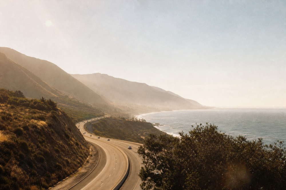 A coastal freeway running north beside the Pacific with hills on one side and the ocean on the other under a hazy California sky, no text, no logos, no people