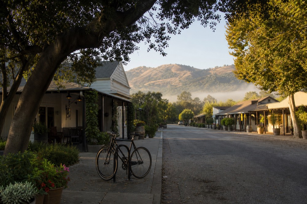 A walkable Los Olivos street of low adobe and clapboard tasting rooms shaded by trees with parked bicycles and flower planters, no text, no logos, no people