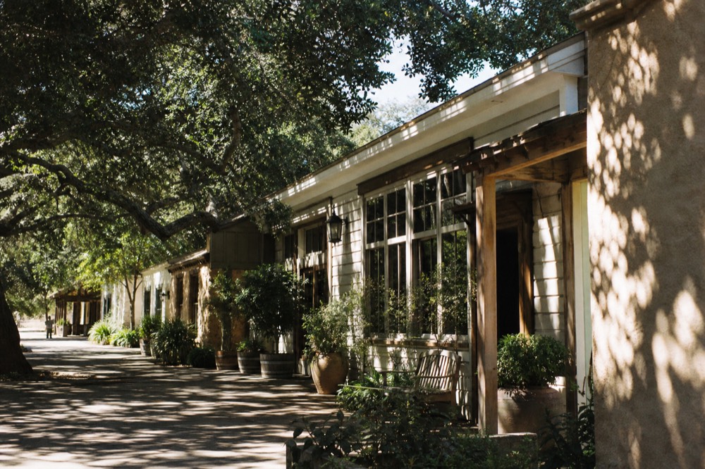 A row of small clapboard and adobe tasting-room storefronts along a Los Olivos village street shaded by oak trees, no text, no logos, no people