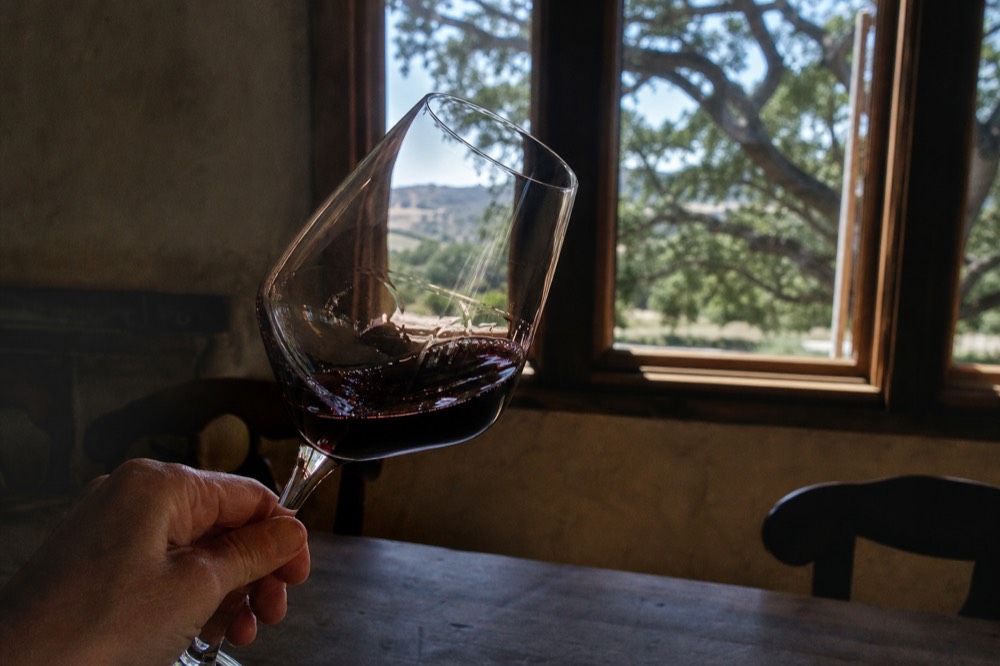 A hand-labeled red wine swirling in a glass held up to soft window light in a rustic tasting room, no text, no logos, no people