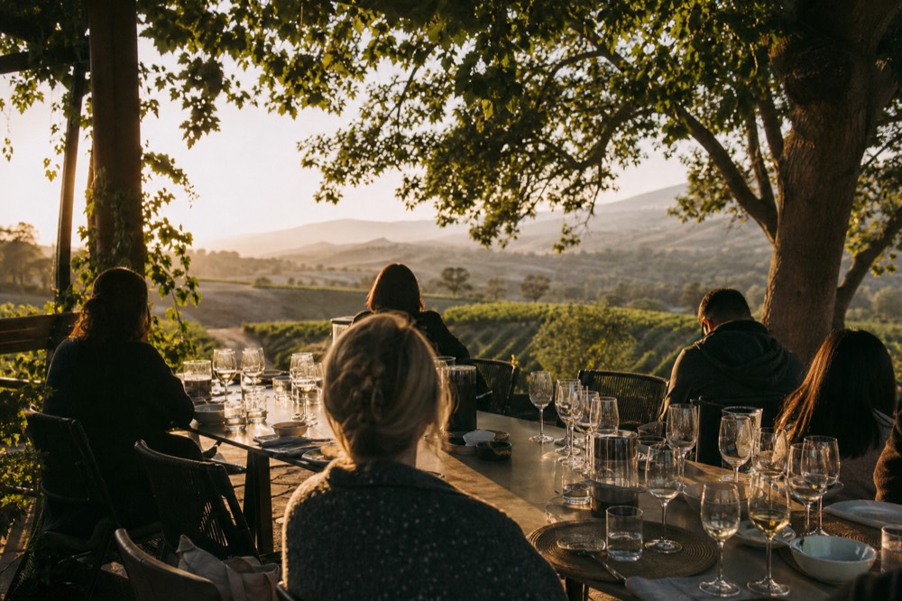 A small group seated at a long private table on a vineyard terrace with tasting glasses and an open valley view behind, warm light, no text, no logos, no people