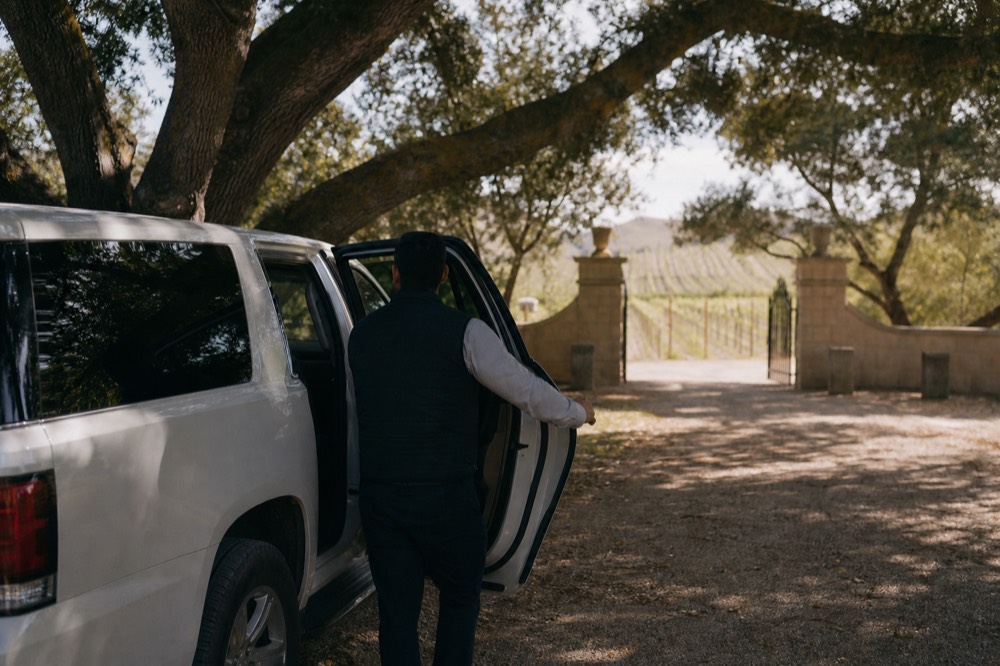 A driver-guide holding open the door of a clean touring vehicle outside a vineyard estate gate with rows of vines beyond, no text, no logos, no people