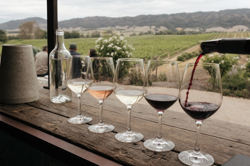 A tasting flight of several wine glasses lined up on a rustic wooden counter with a pour in progress, warm light, no text, no logos, no people