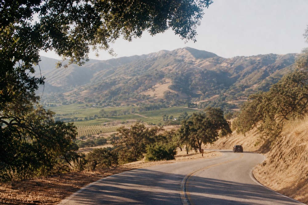 A winding two-lane road climbing over a mountain pass with oak-studded golden hills and a glimpse of vineyards in the valley below, no text, no logos, no people