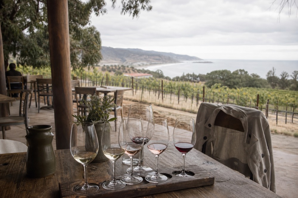 A rustic wooden table on a shaded vineyard patio set with a tasting flight of glasses and a light jacket draped over a chair, vines beyond, no text, no logos, no people