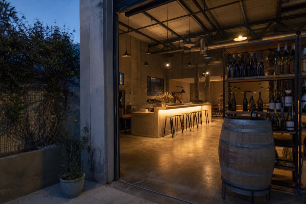 A converted warehouse tasting room in the Funk Zone with an open roll-up door, a concrete-floored bar and wine bottles on shelves, warm afternoon light, no text, no logos, no people