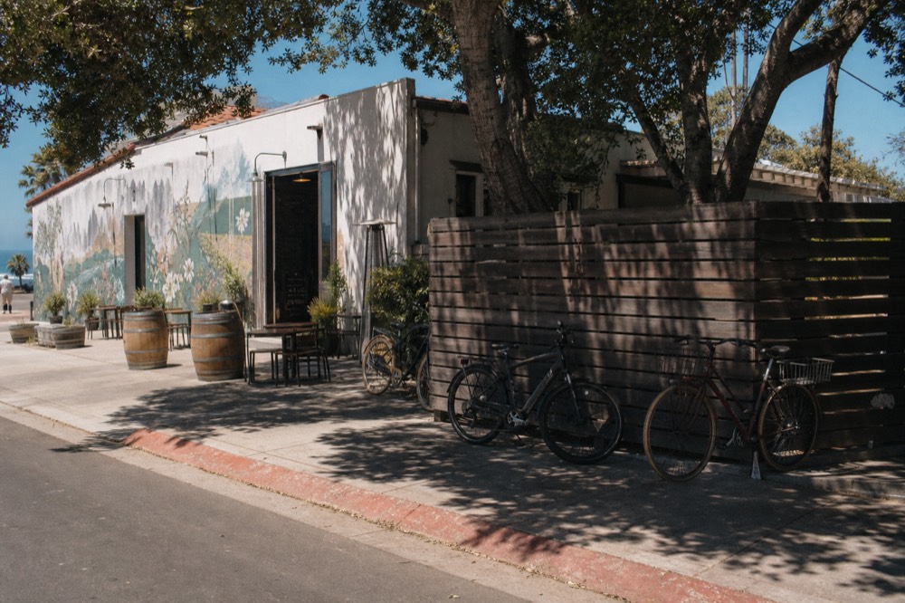 A flat downtown street corner near the Santa Barbara waterfront with a tasting room, a mural-covered wall and bicycles leaning against a fence, no text, no logos, no people