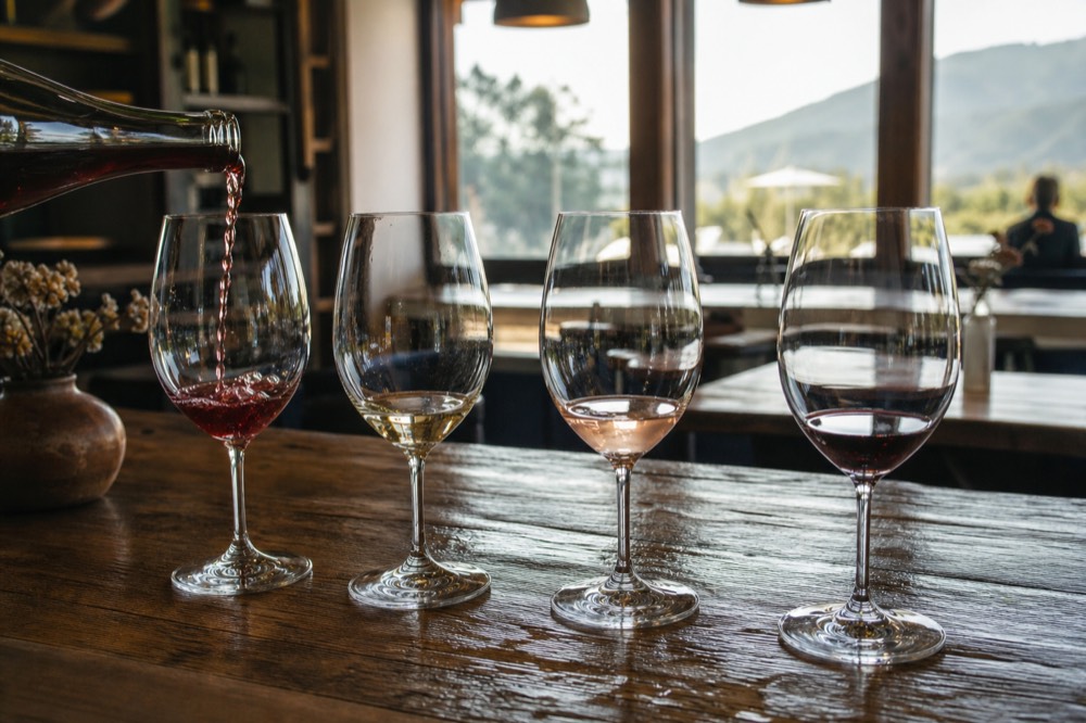 A tasting flight of four wine glasses lined up on a weathered wooden bar top with a pour in progress, warm window light behind, no text, no logos, no people