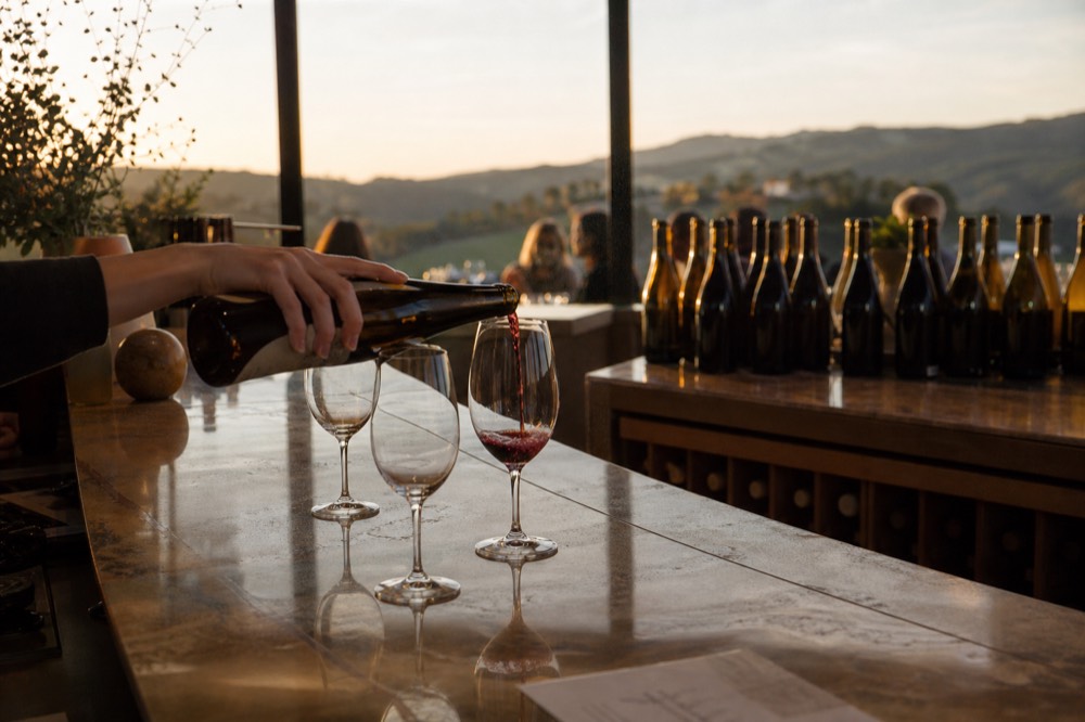 A host pouring a red wine into a stemmed glass across a polished tasting bar with bottles arranged behind in warm light, no text, no logos, no people