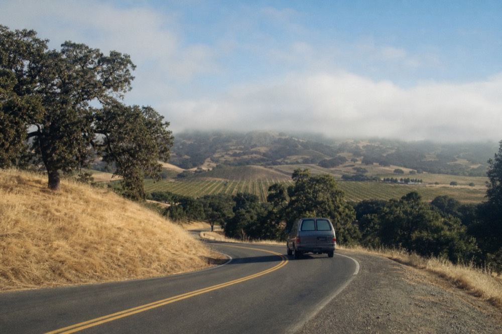 A small touring van on a winding road climbing the San Marcos Pass with oak-studded hills and vineyards below, no text, no logos, no people