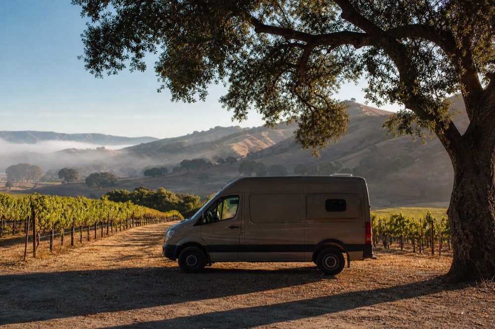 A small touring van parked beside oak-shaded vineyard rows on a Santa Ynez Valley ranch road in warm afternoon light, no text, no logos, no people