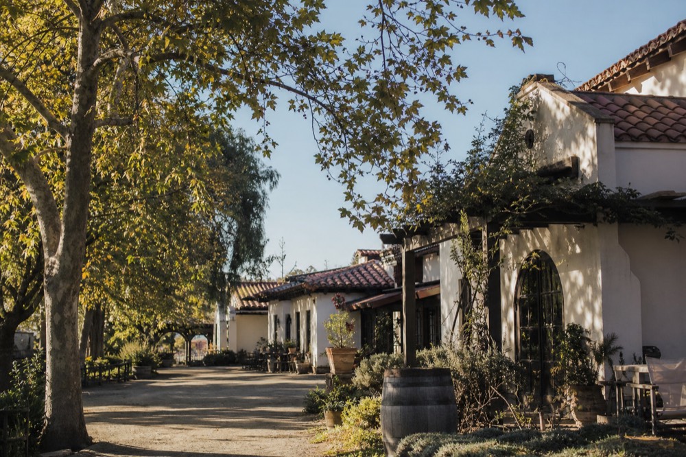 A cluster of low Spanish-colonial adobe and red-tile tasting-room buildings along a quiet Los Olivos street shaded by trees, no text, no logos, no people