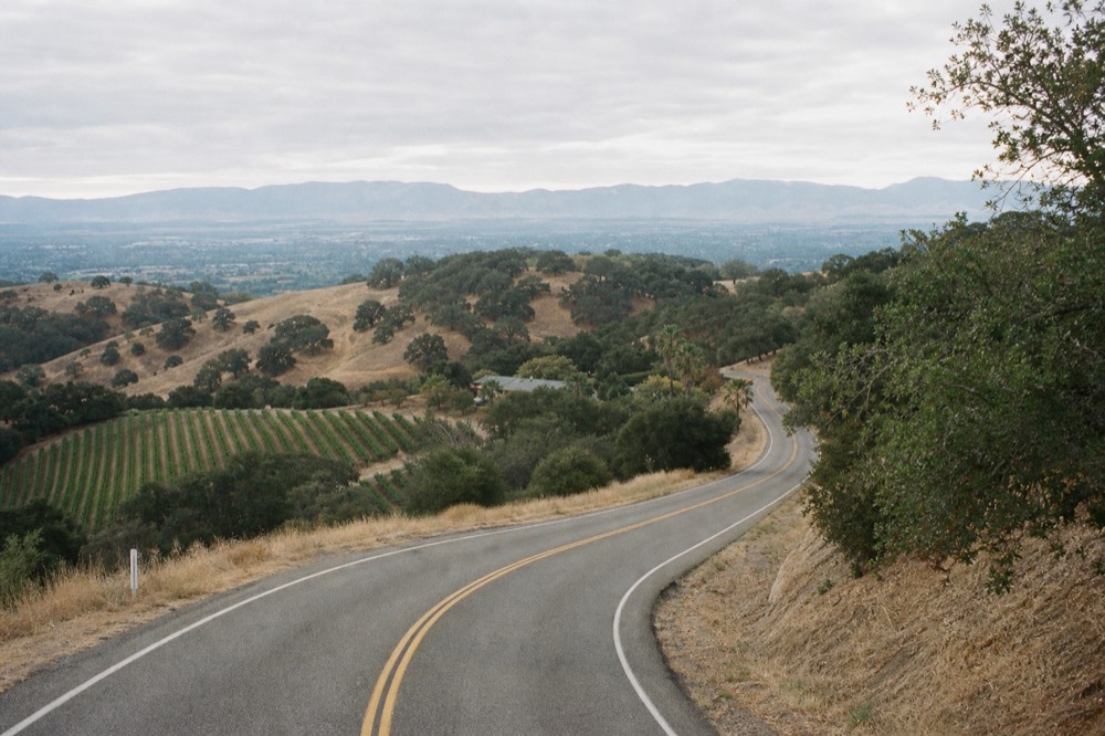 A winding two-lane road climbing over the San Marcos Pass with oak-studded golden hills and a glimpse of vineyards below, no text, no logos, no people