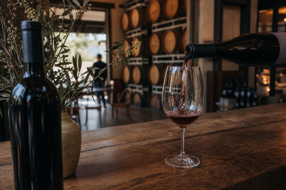 A wine pour caught mid-stream into a glass at a rustic cellar-door counter with barrels blurred behind, warm interior light, no text, no logos, no people