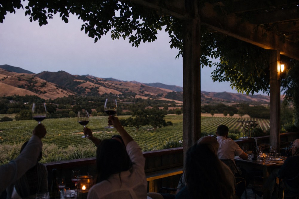 Guests raising glasses on a shaded terrace overlooking Santa Ynez vineyard rows and golden hills at the end of a tasting day, no text, no logos, no people